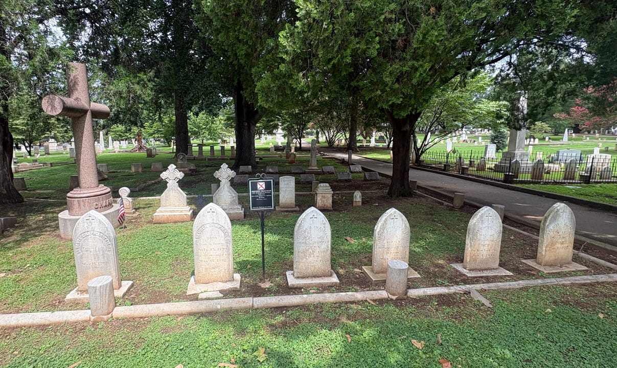A row of 19th-century headstones and a carved cross monument along a tree-lined section of Maple Hill Cemetery, Huntsville, Alabama.