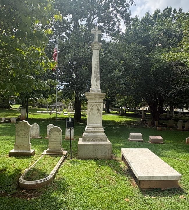 A tall cross-topped monument and flagpole among headstones under mature trees at Maple Hill Cemetery, Huntsville, Alabama.