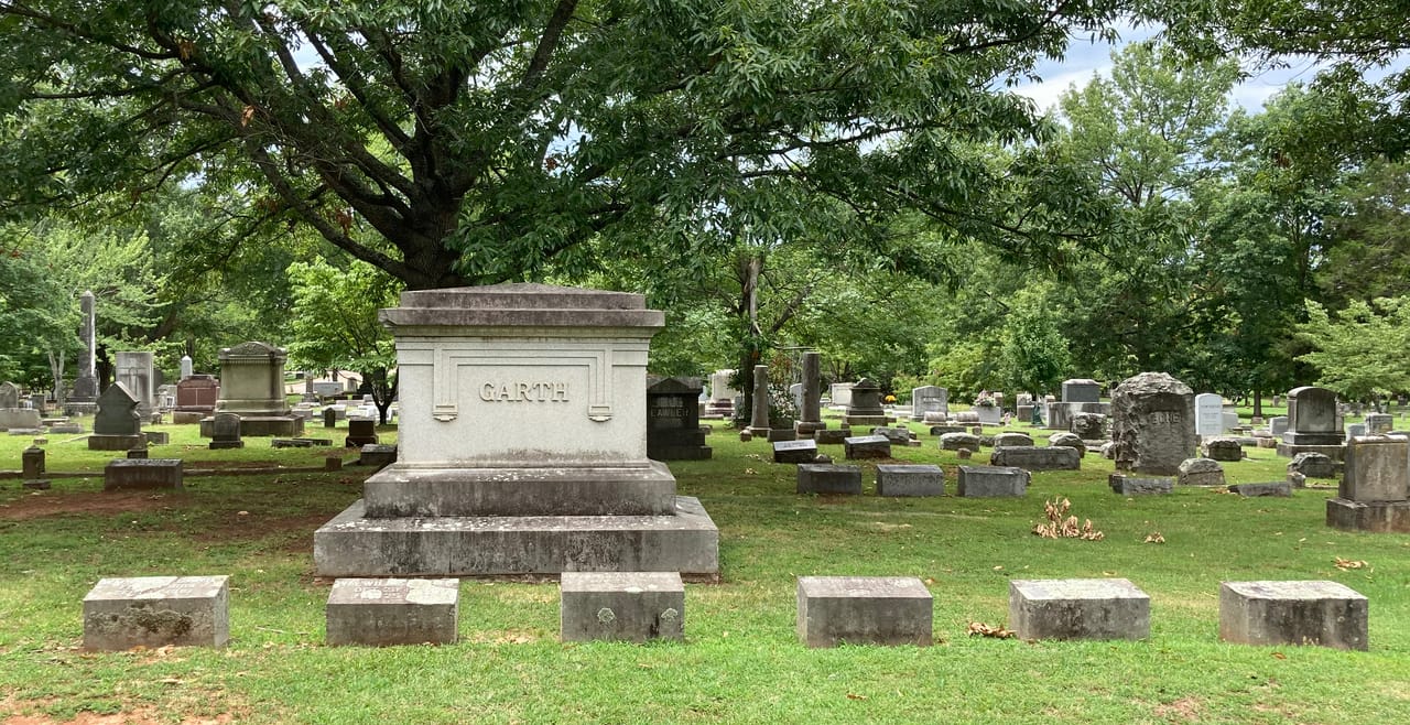 A wide view of a stone family monument and rows of surrounding headstones beneath a canopy of mature trees at Maple Hill Cemetery, Huntsville, Alabama.