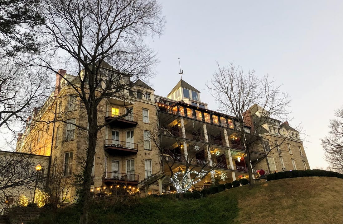 The Crescent Hotel's stone wing and tiered porches on the hillside at dusk, decorated with holiday lights, seen through bare trees, Eureka Springs, Arkansas, December 2021.