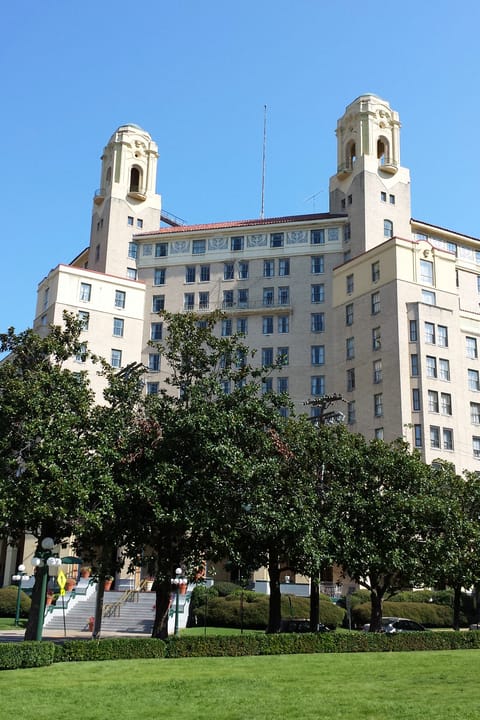 A vertical front-on view of the full Arlington Hotel building with its twin towers and ground-floor arcade, framed by lawn and trees.