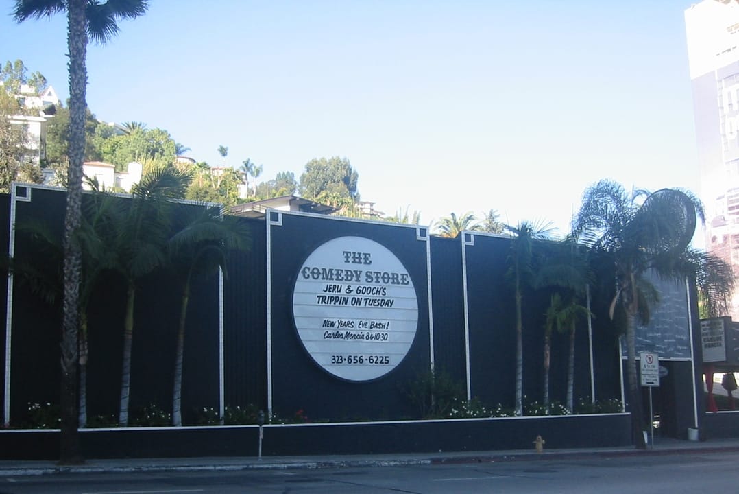 Close view of The Comedy Store's wall-mounted sign and hours plaque along Sunset Boulevard, with the row of palm trees in front.