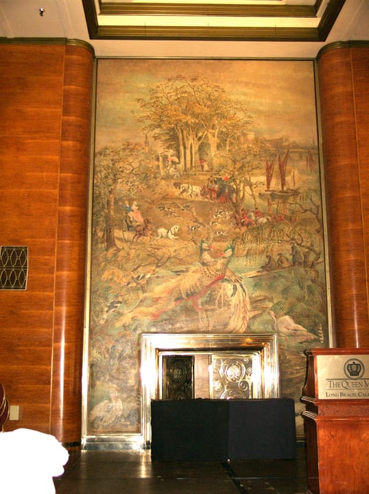 A large painted mural and fireplace in one of the Queen Mary's grand public rooms, beside a lectern lettered "The Queen Mary, Long Beach, CA".