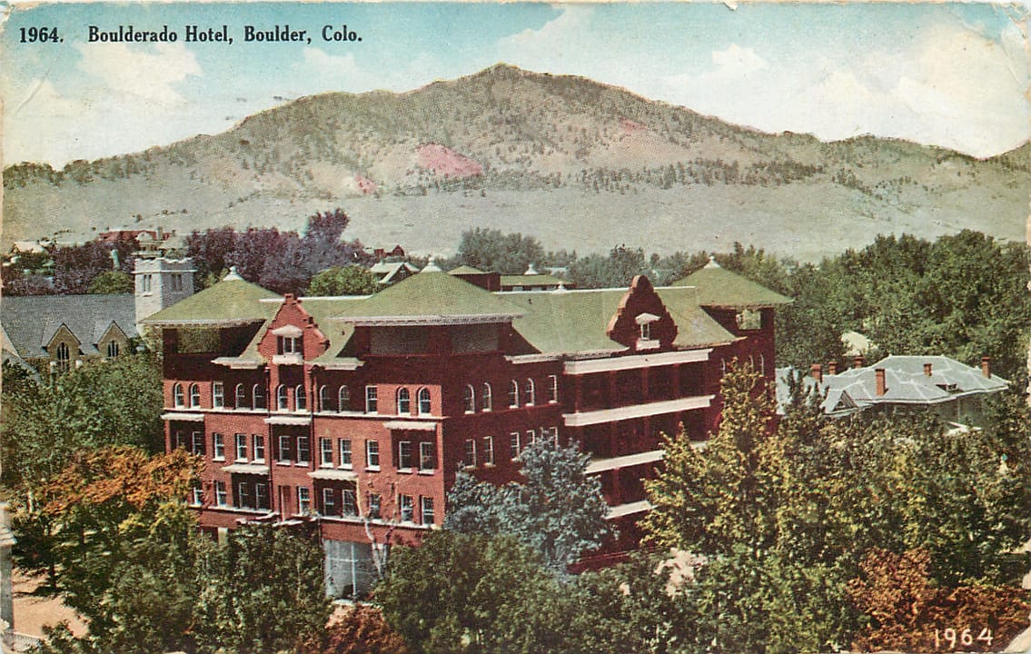 The Hotel Boulderado's red-brick, Spanish Renaissance-style facade on 13th Street in downtown Boulder, Colorado, with state and national flags flying from the second-floor balcony.