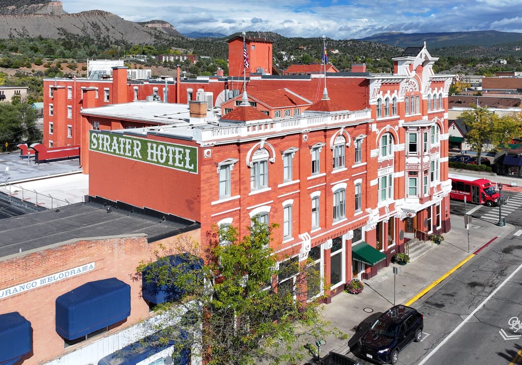 An elevated view of the Strater Hotel in downtown Durango, Colorado, with the 'STRATER HOTEL' roof sign visible and the surrounding mountains in the background.
