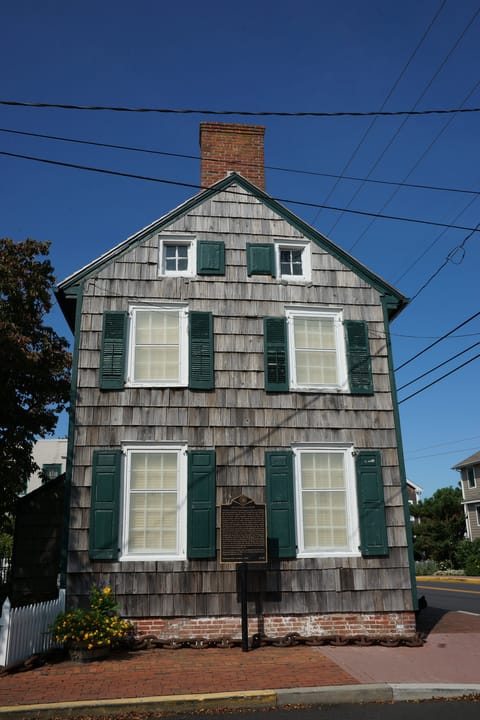 The gable end of the Cannonball House on Front Street in Lewes, Delaware: shingled walls, green shutters, a brick chimney and a state historical marker beside the lower windows.