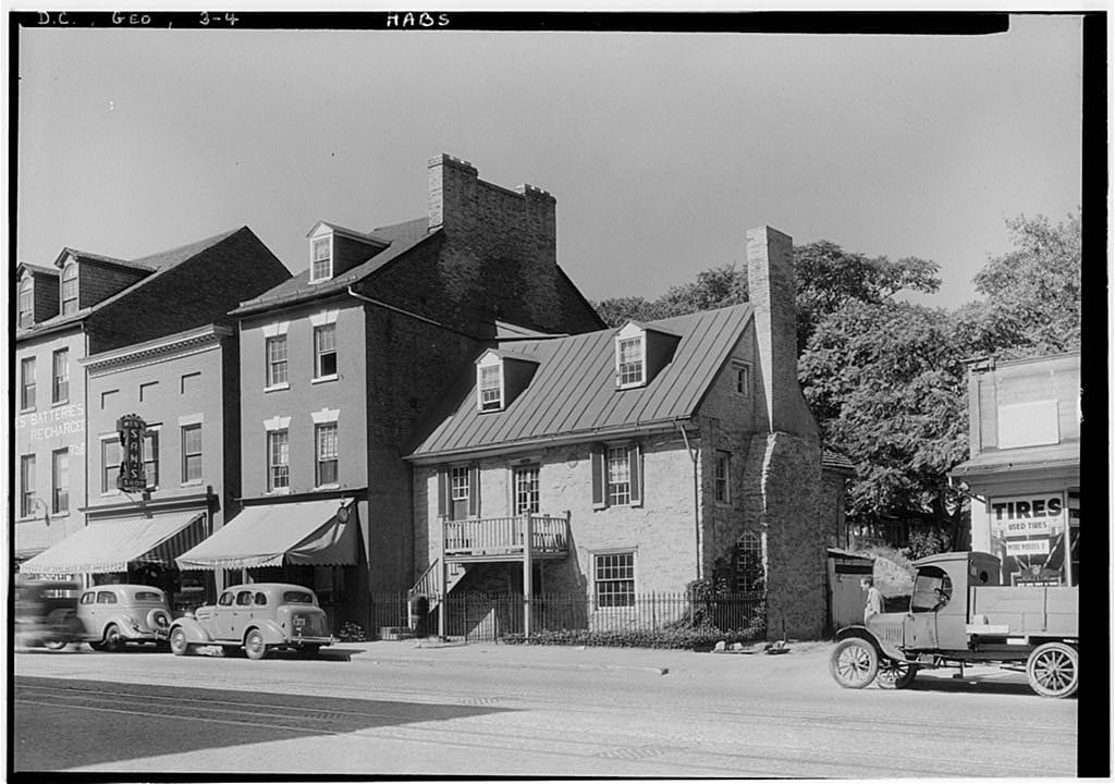 The Old Stone House on M Street in Georgetown, seen from the southeast in October 1935, beside brick commercial buildings, in a Historic American Buildings Survey photograph by Albert S. Burns.