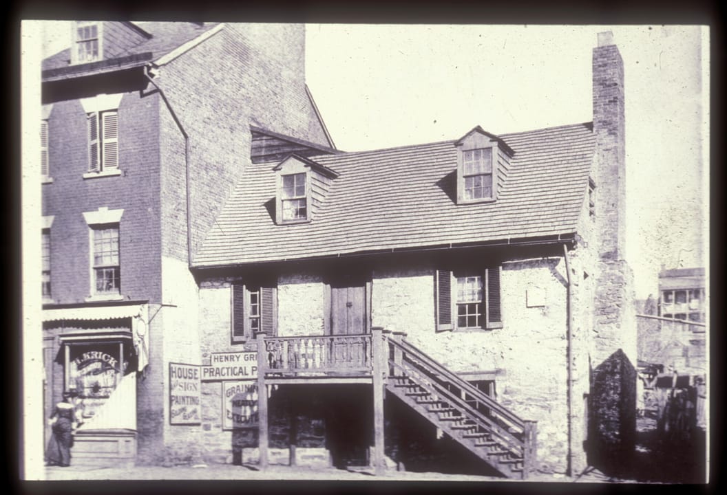 The Old Stone House in Georgetown in the late 1800s, with a wooden stair running down from the second-floor door on the east side and painters' signs on the front wall.