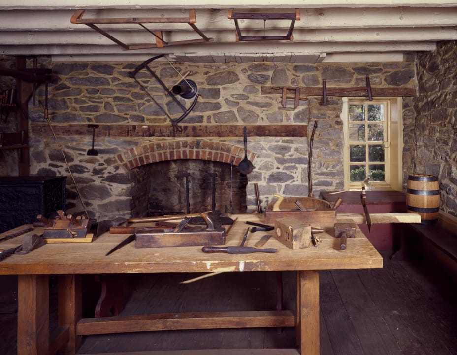 The cabinetmaker's workshop room on the ground floor of the Old Stone House in Georgetown, with a stone wall, a brick-arched fireplace, a workbench and woodworking tools.
