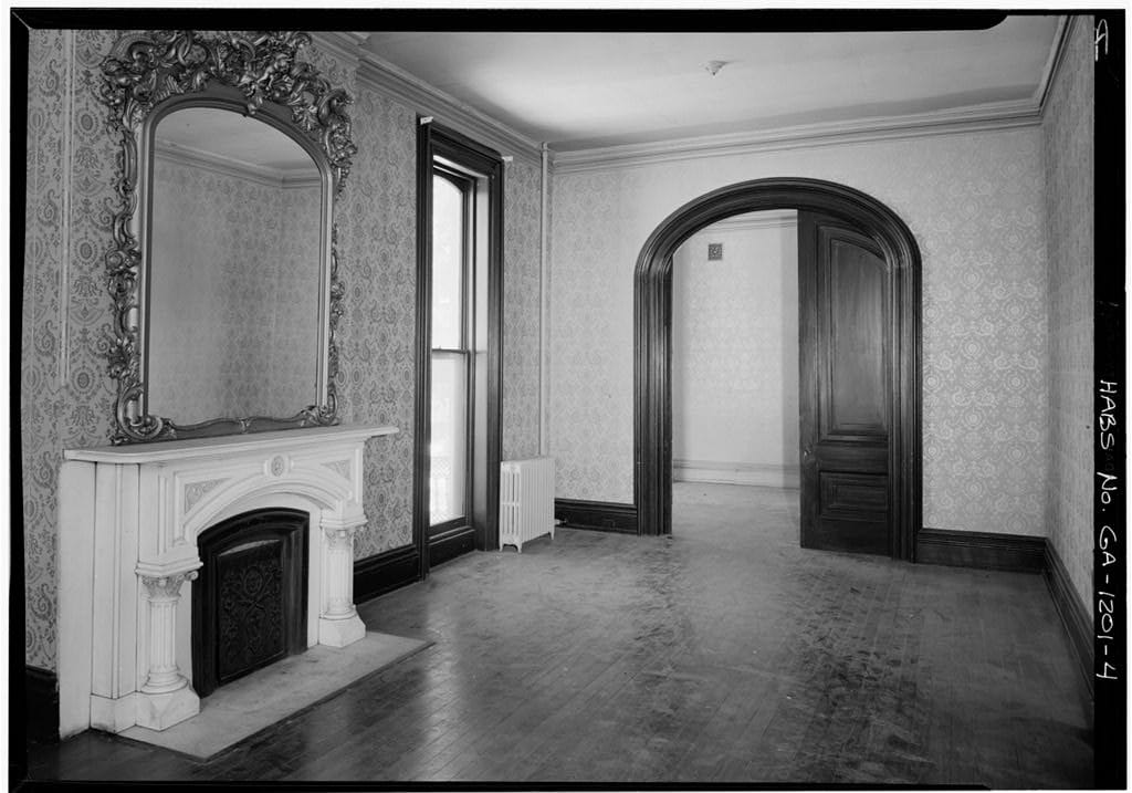 The empty first-floor ballroom of the Hamilton-Turner house in Savannah in a June 1966 Historic American Buildings Survey photograph, with a marble fireplace, a tall mirror, patterned wallpaper and an arched doorway.