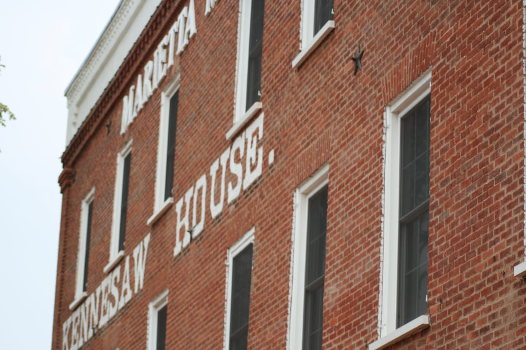 Part of the red-brick wall of the Kennesaw House in Marietta, Georgia, seen from below at an angle, with window surrounds, a white cornice, star-shaped wall anchors and the building's painted white lettering.