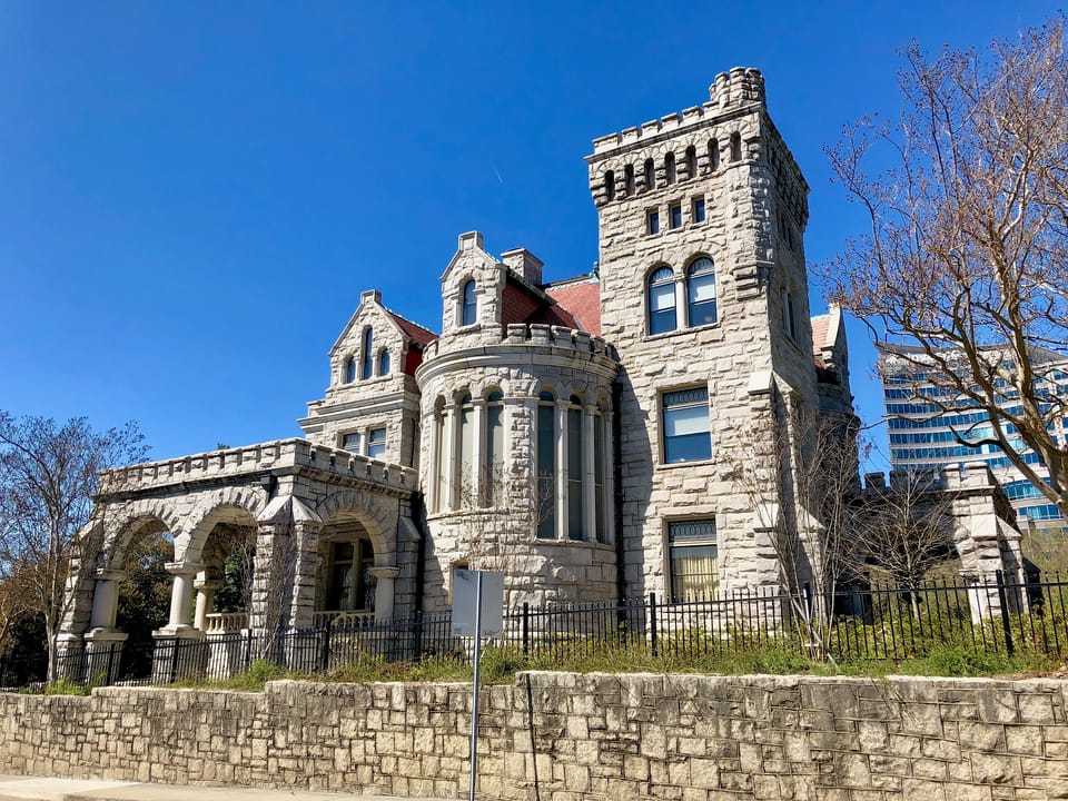 Corner view of Rhodes Hall, the 1904 Romanesque Revival granite mansion on Peachtree Street in Atlanta, showing the crenellated tower, round turret and arched porch above the rubble retaining wall, March 2019.