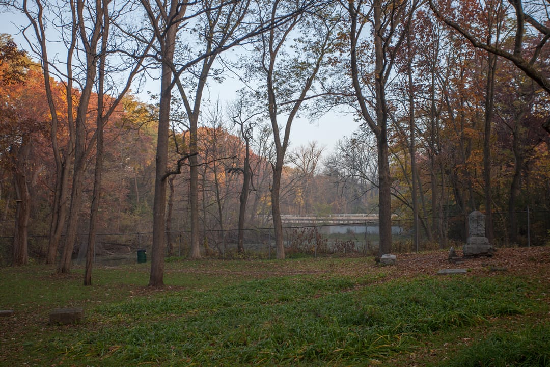 A wide view of the wooded clearing at Bachelor's Grove Cemetery, with scattered headstones and the pond visible in the distance, autumn afternoon.