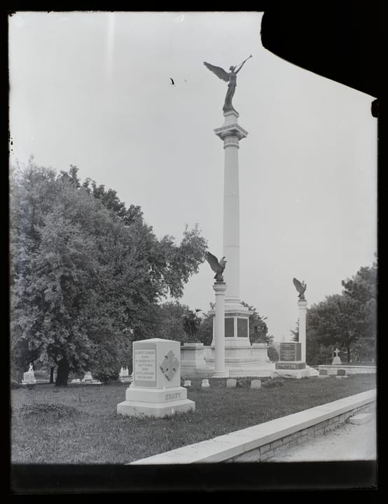 Vertical, black and white photograph showing the Elijah Parish Lovejoy monument in Alton City Cemetery in Alton, Illinois. The monument has a tall pillar topped with an angel blowing a trumpet. The tall pillar is flanked by two smaller pillars that are topped with eagles. Other gravestones surround 