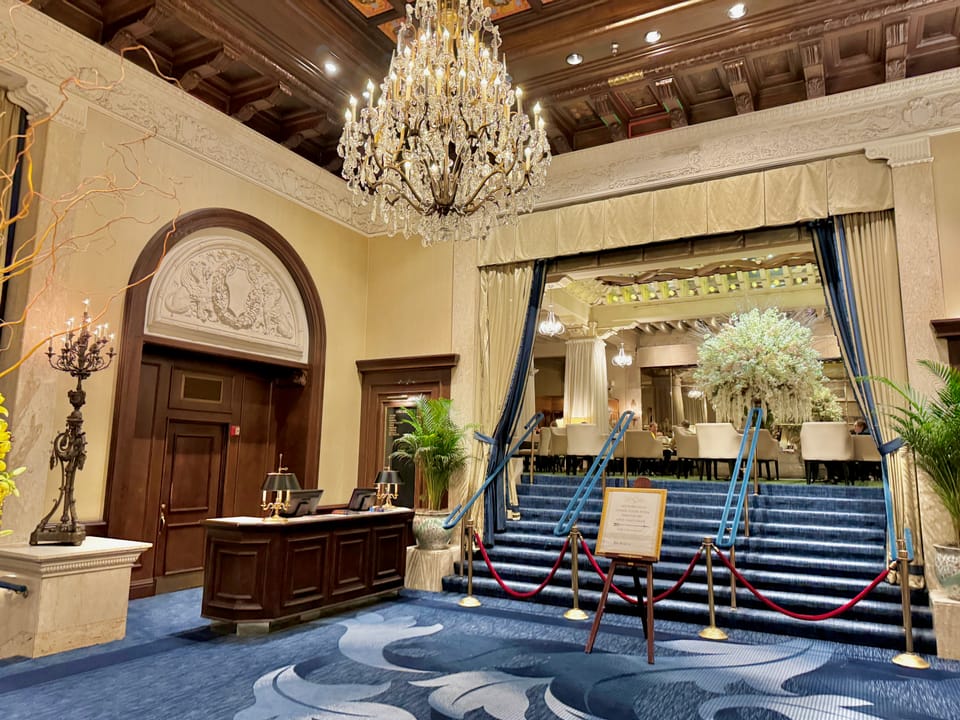 The public lobby of the Drake Hotel, with a crystal chandelier, coffered wood ceiling, a desk and a carpeted stair behind rope stanchions, Chicago, October 2024.