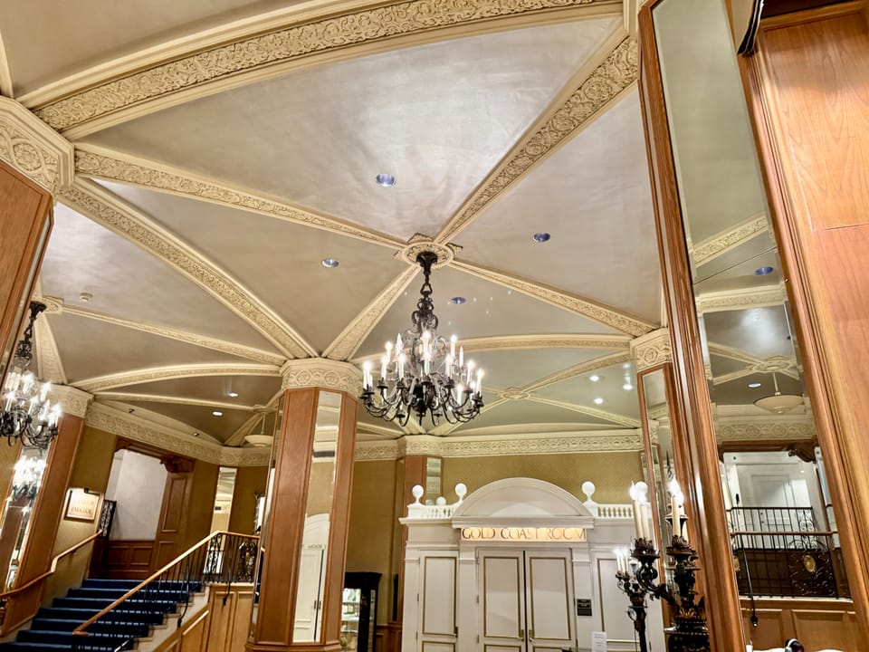 The ornamented ceiling and chandelier of a public lobby landing at the Drake Hotel, with a stair at the left and the doors lettered 'Gold Coast Room' at the right, Chicago, October 2024.