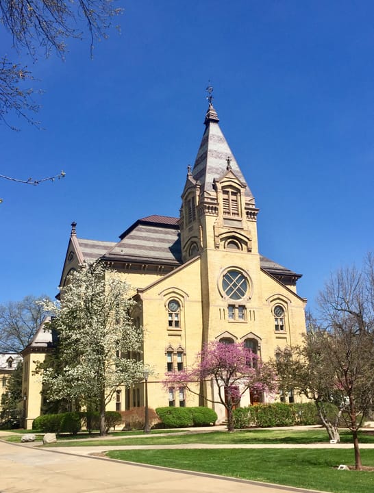 A closer view of Washington Hall's taller tower, with its dark conical roof, weathervane finial and arched window, at the University of Notre Dame.