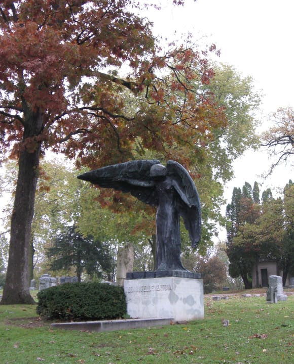 Full view of the Black Angel monument on its granite base at Oakland Cemetery, Iowa City, Iowa, with surrounding grave markers and trees in fall color.