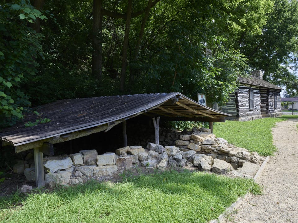 A stone-walled well or root cellar with a wood lean-to roof on the grounds of the Mathias Ham House Historic Site, Dubuque, Iowa; the log cabin is visible behind it.