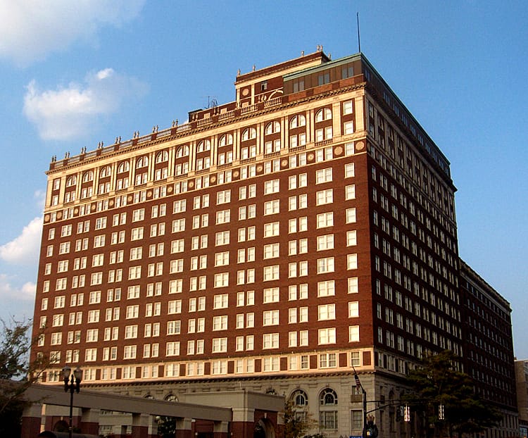 Street-level corner view of The Brown Hotel's full Georgian Revival brick facade at 4th Street and Broadway in downtown Louisville, with the hotel's sidewalk clock and red awnings.