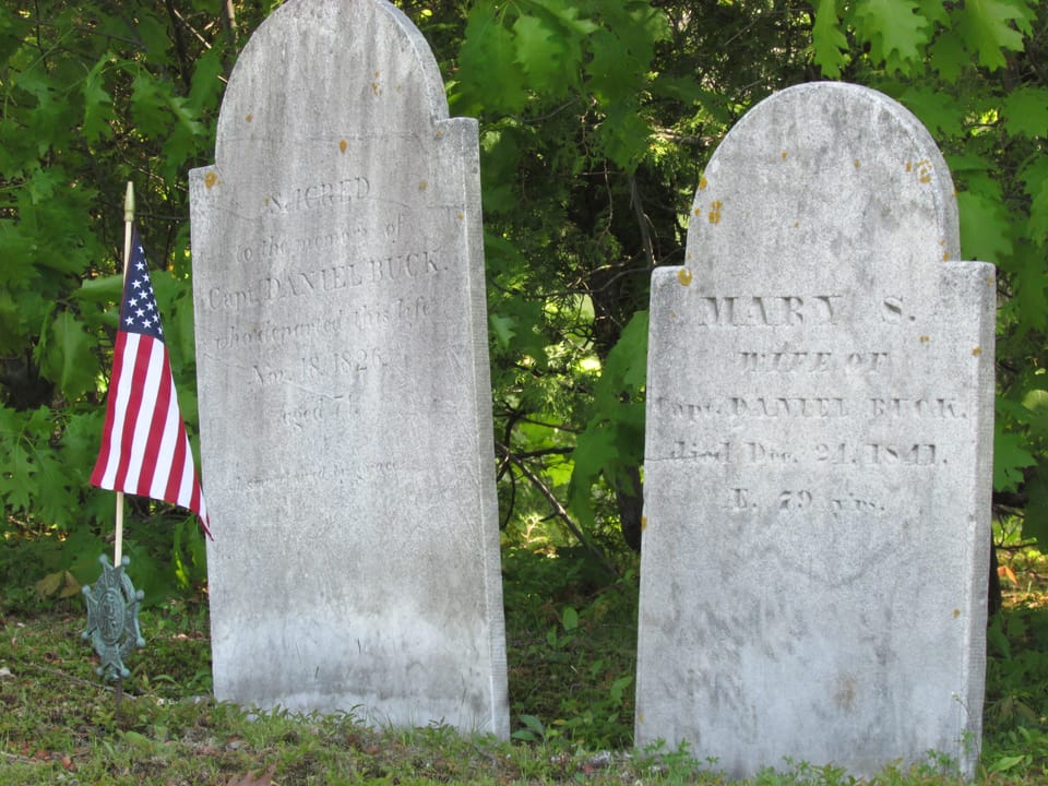 The weathered 19th-century headstones of Capt. Daniel Buck (died 1826) and his wife Mary (died 1841), with a small flag and a veterans' grave marker, in Buck Cemetery, Bucksport, Maine.