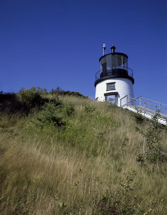Owls Head Light seen from the grassy hillside below, looking up at the tower and its approach stairway under a clear blue sky.