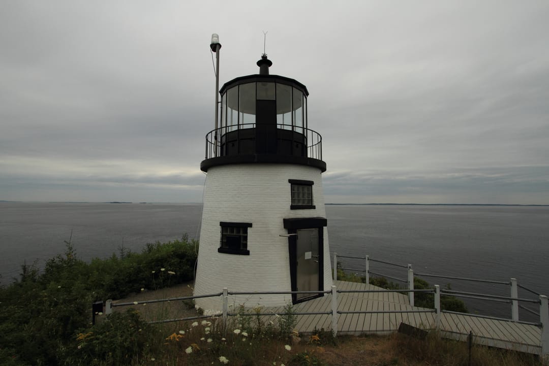 Owls Head Light under an overcast sky with the open ocean behind it, seen from the boardwalk at the base of the tower.