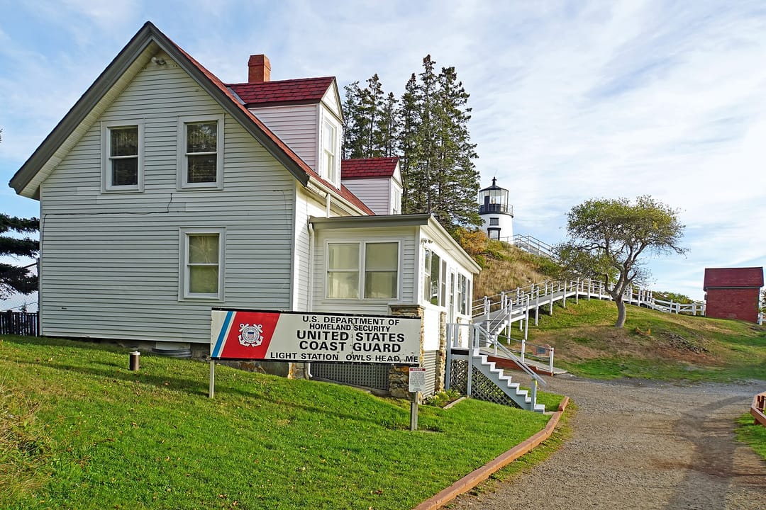 The keeper's house at Owls Head Light Station, with the lighthouse tower visible behind it and the U.S. Coast Guard station sign ('United States Coast Guard Light Station Owls Head, ME') in the foreground.