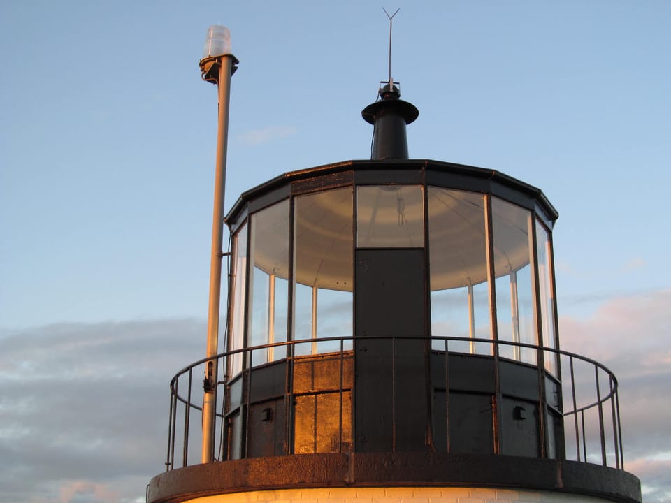 Close detail of the lighthouse's lantern room and lens apparatus at Owls Head Light, with its brass fixtures lit against the evening sky.