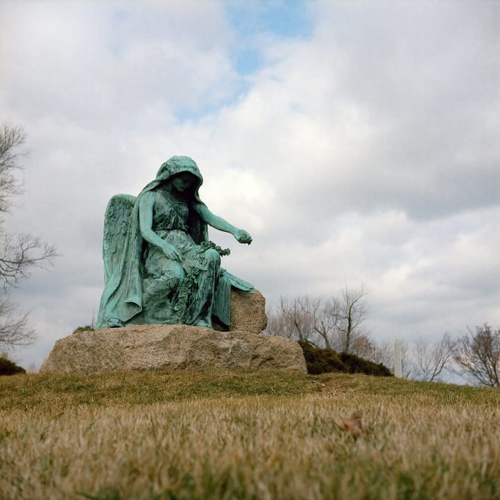 The Clotho statue at Druid Ridge Cemetery, Pikesville, Maryland: a bronze hooded, winged figure seated on a granite boulder with a wreath across her lap and one arm extended, seen from a low angle under a winter sky. A separate monument from Black Ag