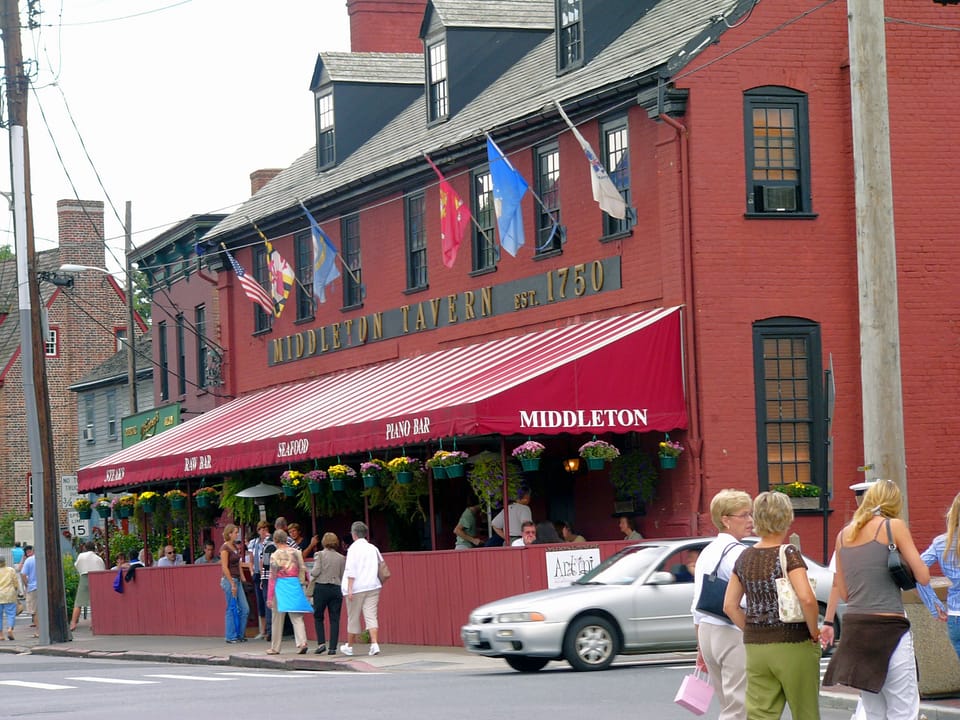 Middleton Tavern's red-brick building at the Annapolis waterfront, with its dormered roof, flags, 'Middleton Tavern Est. 1750' lettering and striped awning over the outdoor seating, September 2005.
