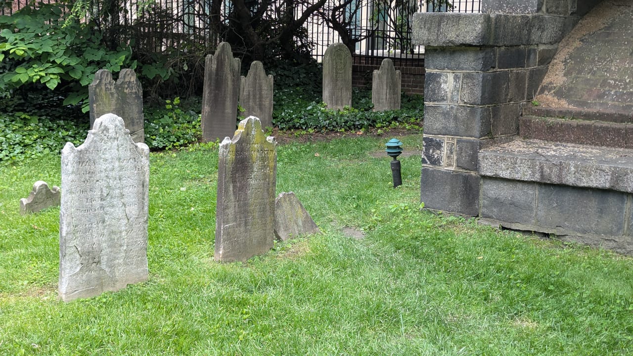 A landscape view inside Westminster Burying Ground, with period headstones on the lawn and a corner of the brick churchyard wall