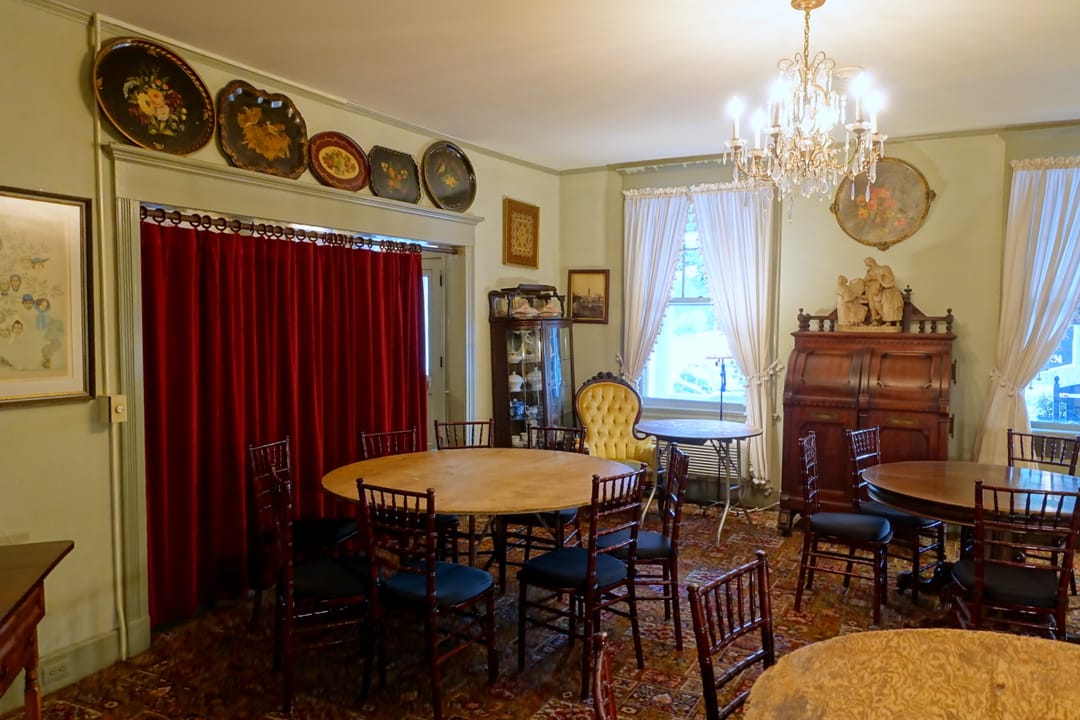 A dining room at the Red Lion Inn, with a chandelier, round tables, red velvet curtains and painted trays hung on the wall, Stockbridge, Massachusetts, 2017.