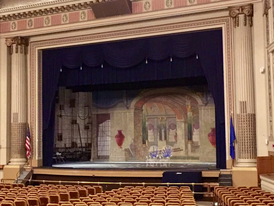 The auditorium stage and proscenium arch, seen from the orchestra seats, flanked by the US and Minnesota state flags and painted murals.