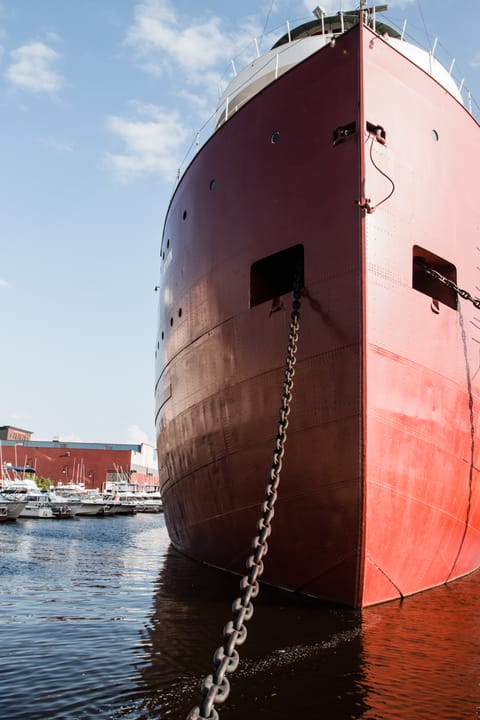 Dramatic bow-on view of the SS William A. Irvin's prow and anchor chain at its Duluth berth, with the harbor and nearby buildings visible beyond.