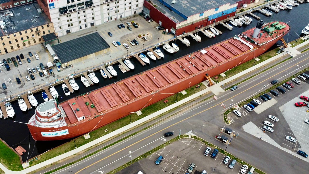 Aerial view of the SS William A. Irvin moored along Harbor Drive at Duluth's Canal Park, with a marina beside it and brick and warehouse buildings behind; the museum ship's bow carries a 'Tour this ship' banner. August 2024.