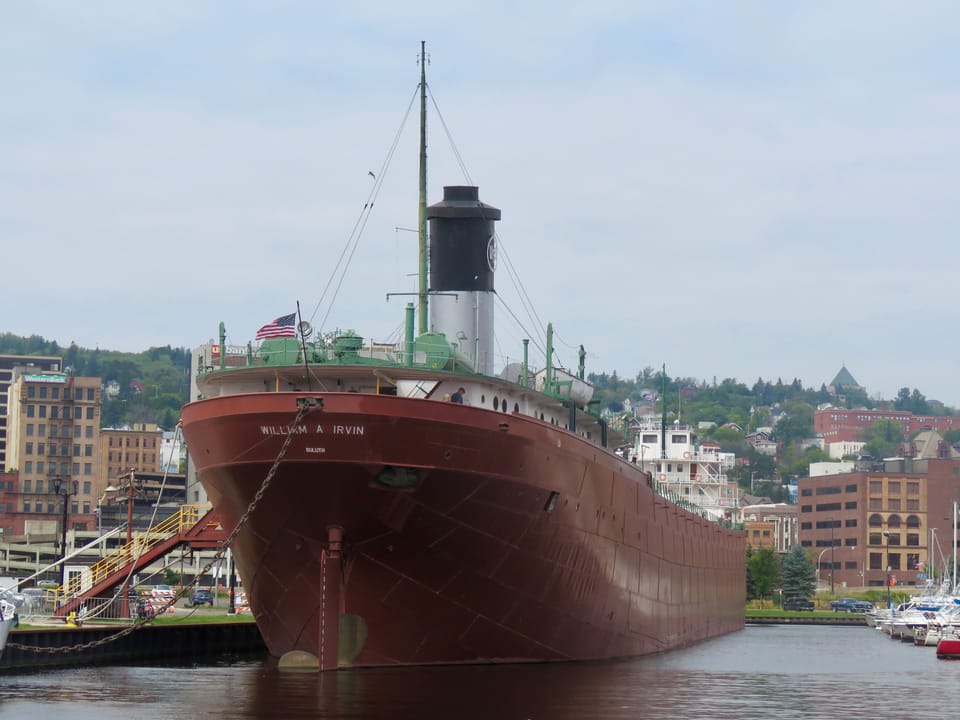 The stern of the SS William A. Irvin seen across the water at its Duluth berth, with 'WILLIAM A IRVIN, DULUTH' on the hull, the aft deckhouse, the stack with the U.S. Steel emblem and the mast above, and Duluth's hillside behind. August 2024.
