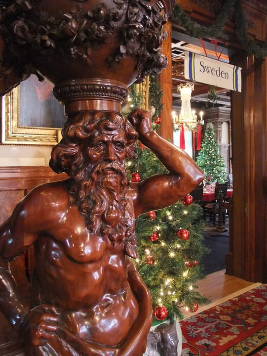 A carved wooden Atlas/Neptune figure in the mansion's interior, decorated for the Institute's annual "Nordic Christmas" exhibit, with a "Sweden" banner visible behind.