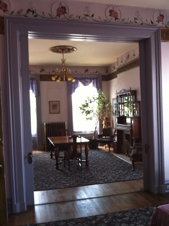 A parlor/dining room inside the Lemp Mansion, viewed through a doorway with period wallpaper and furnishings.