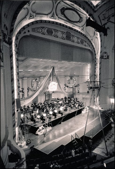 The proscenium arch and stage of Powell Hall's auditorium with an orchestra and holiday wreaths on the rear wall, seen from an upper level (black-and-white film photograph).