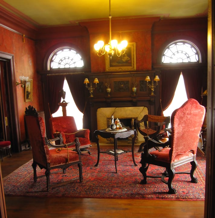 A formal parlor inside Rockcliffe Mansion, with a fireplace, period red upholstered chairs and a chandelier, described by the uploader as 'Mr. Cruikshank's Parlor'.