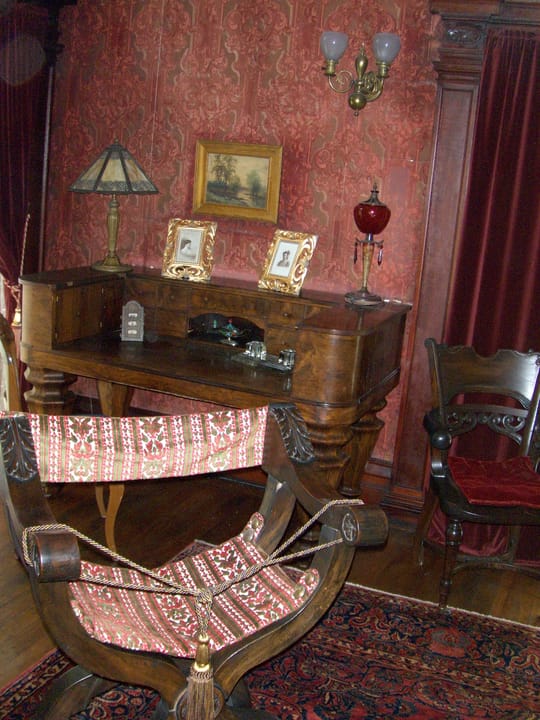 A period writing desk and an upholstered folding chair against patterned red wallpaper inside Rockcliffe Mansion.
