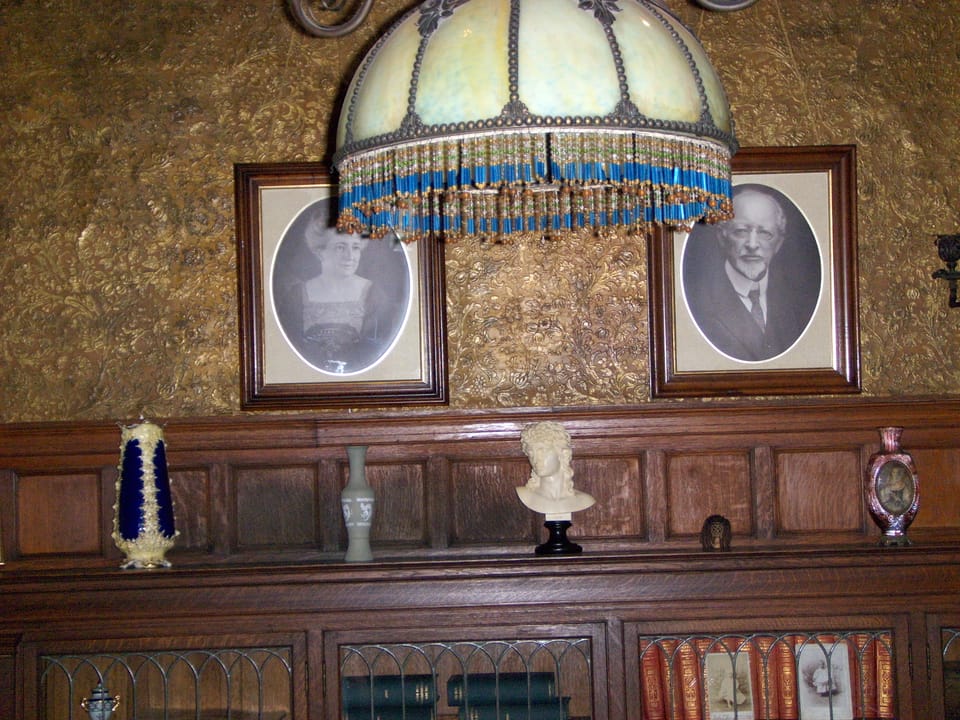 A built-in bookcase inside Rockcliffe Mansion with a stained-glass lamp and framed portrait photographs of the home's former occupants on the shelf.