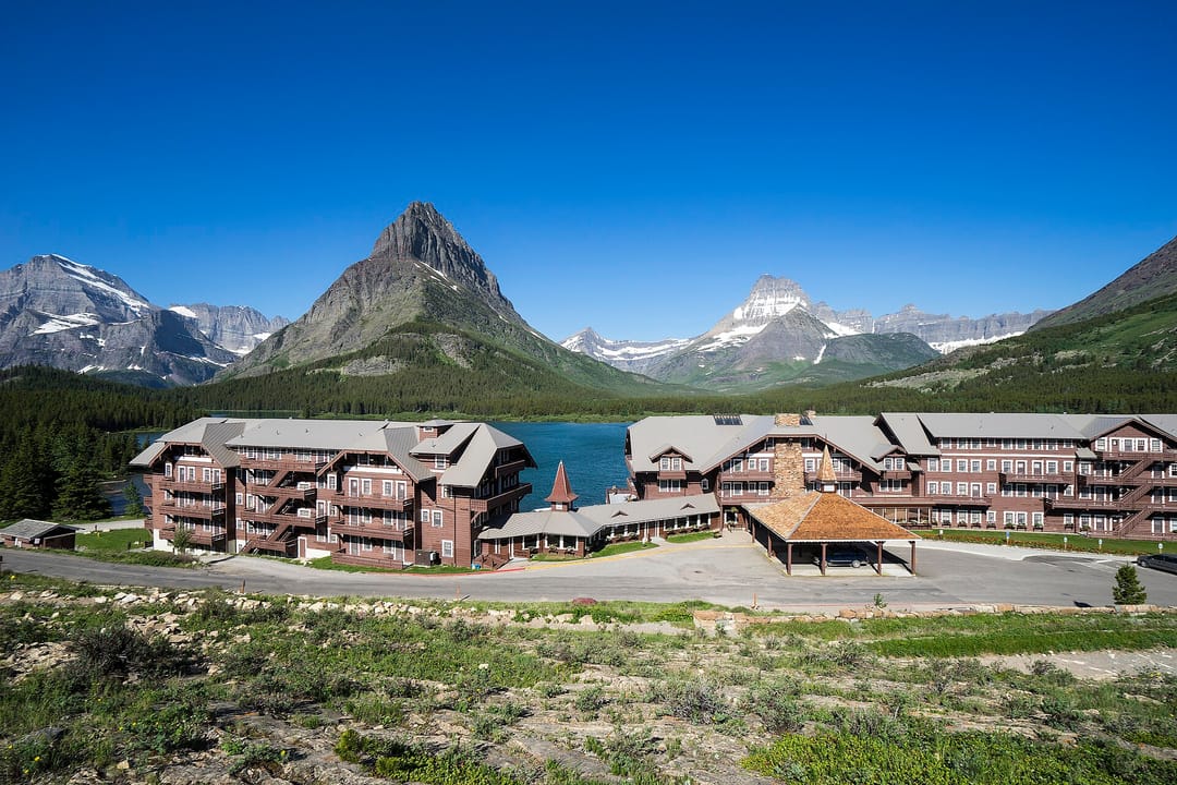 Many Glacier Hotel seen across Swiftcurrent Lake with the Grinnell/Swiftcurrent peaks behind it, a wide daytime view of the full lodge and its wings.