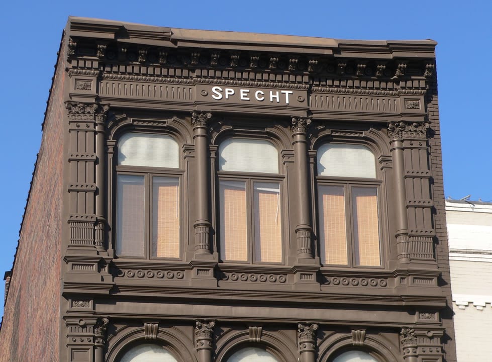 Architectural detail: the upper story of the Christian Specht Building's south facade, ornate pressed-metal window surrounds and cornice with the 'SPECHT' nameplate legible.