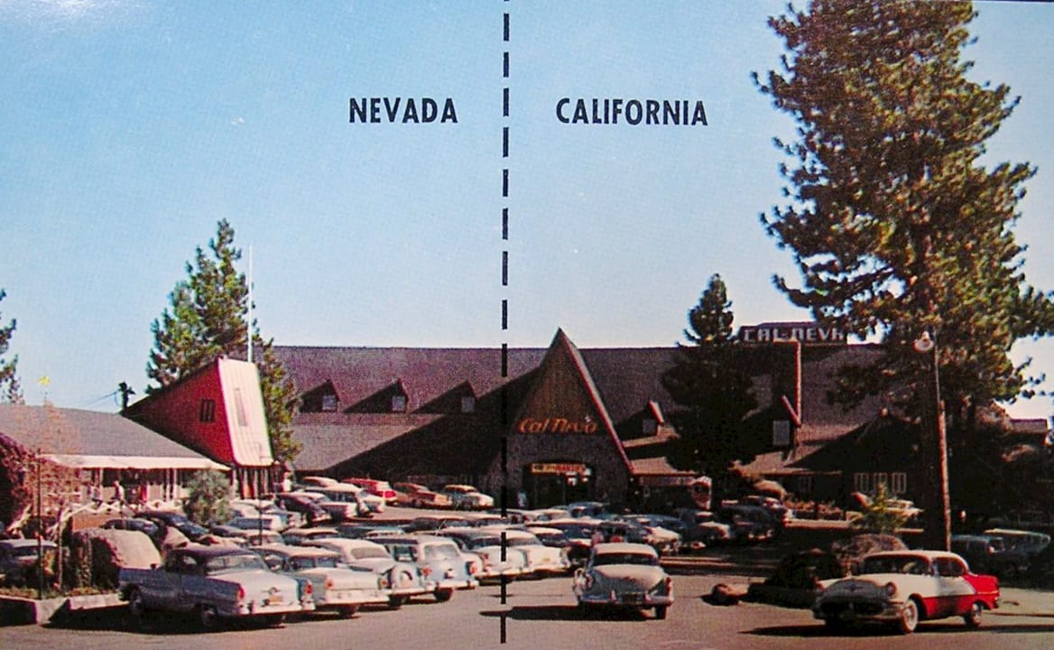 A vintage postcard of the Cal Neva Lodge with the painted 'NEVADA | CALIFORNIA' state line across the building and parking lot full of period cars.