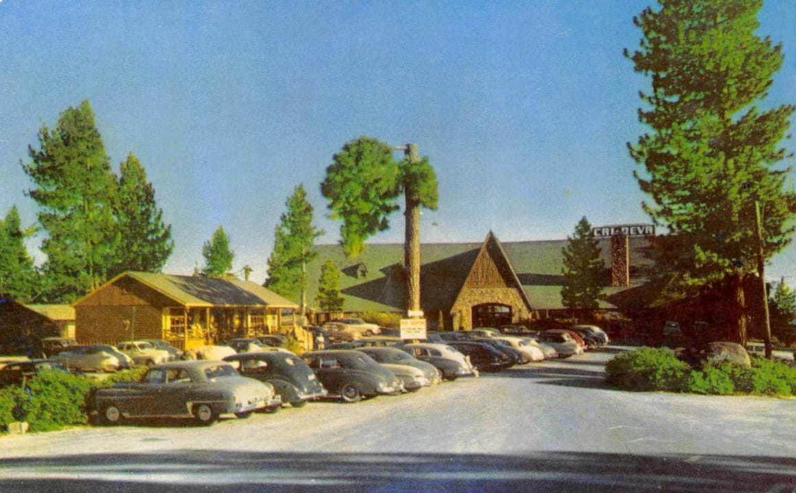 A vintage postcard of the Cal Neva Lodge grounds and parking lot, lined with period cars, among the pines above Lake Tahoe.