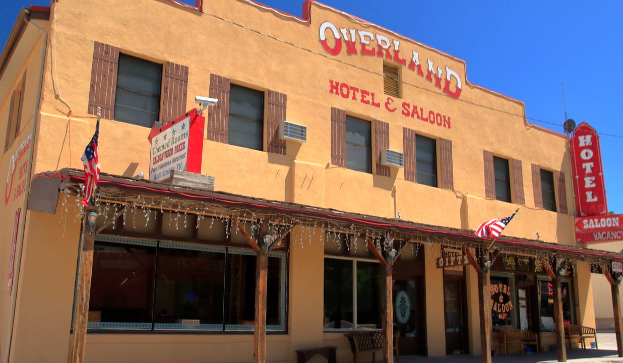 A three-quarter view of the Overland Hotel & Saloon on Main Street, Pioche, Nevada, showing the covered porch, the painted 'Overland Hotel & Saloon' lettering and the red vertical 'HOTEL / SALOON' sign, July 2023.