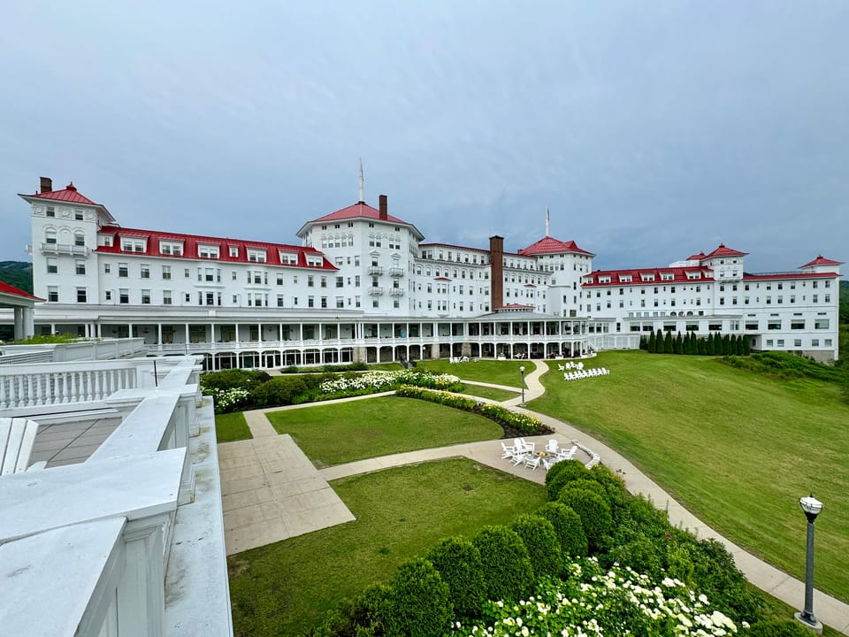The hotel's back lawn and garden paths seen from an upper veranda, Bretton Woods, NH, with the building's red roofline and wraparound porch in the background.