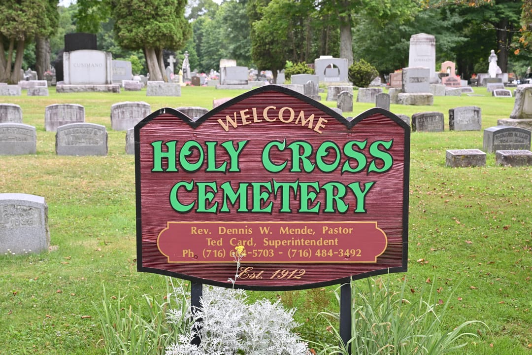 The sign says: "Welcome", "Holy Cross Cemetery", "Rev. Dennis W. Mende, Pastor", Ted Card, Superintendent", "ph (716) 664-5703 - (716) 484-3492", "Est. 1912". 1 West Oak Hill Road, Jamestown, New York, 14701.